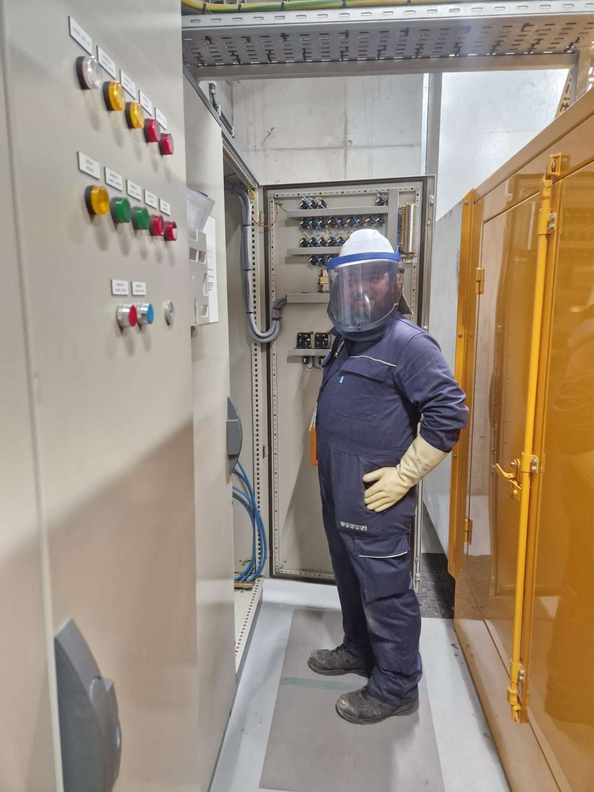 Opérateur en équipement de protection individuelle — casque, écran facial anti-arc et gants isolants — devant une armoire électrique basse tension ouverte.